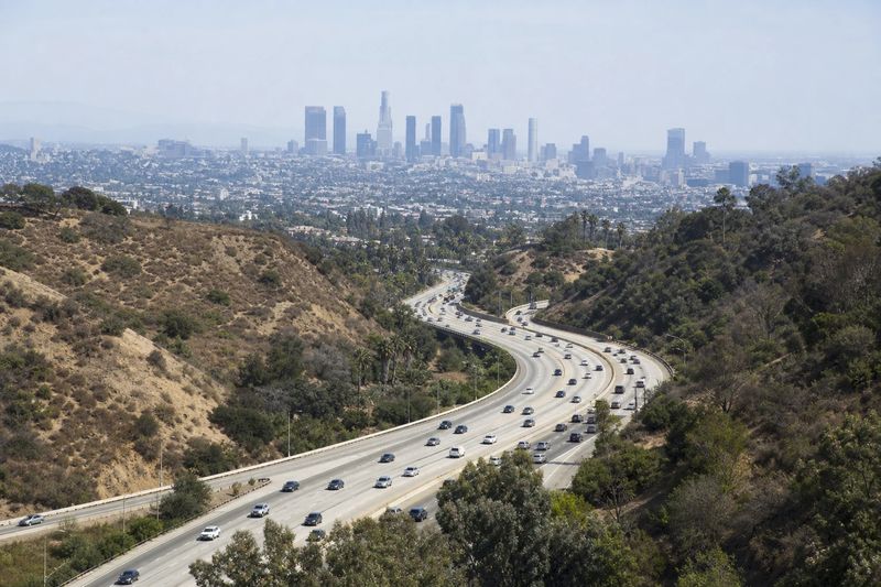 A multi-lane Los Angeles freeway curving toward downtown towers with hills in the distance